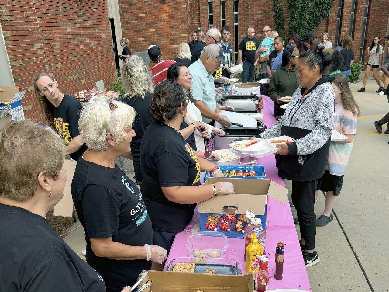 Volunteers serving food to neighbors along a line of tables at the Free Neighborhood BBQ