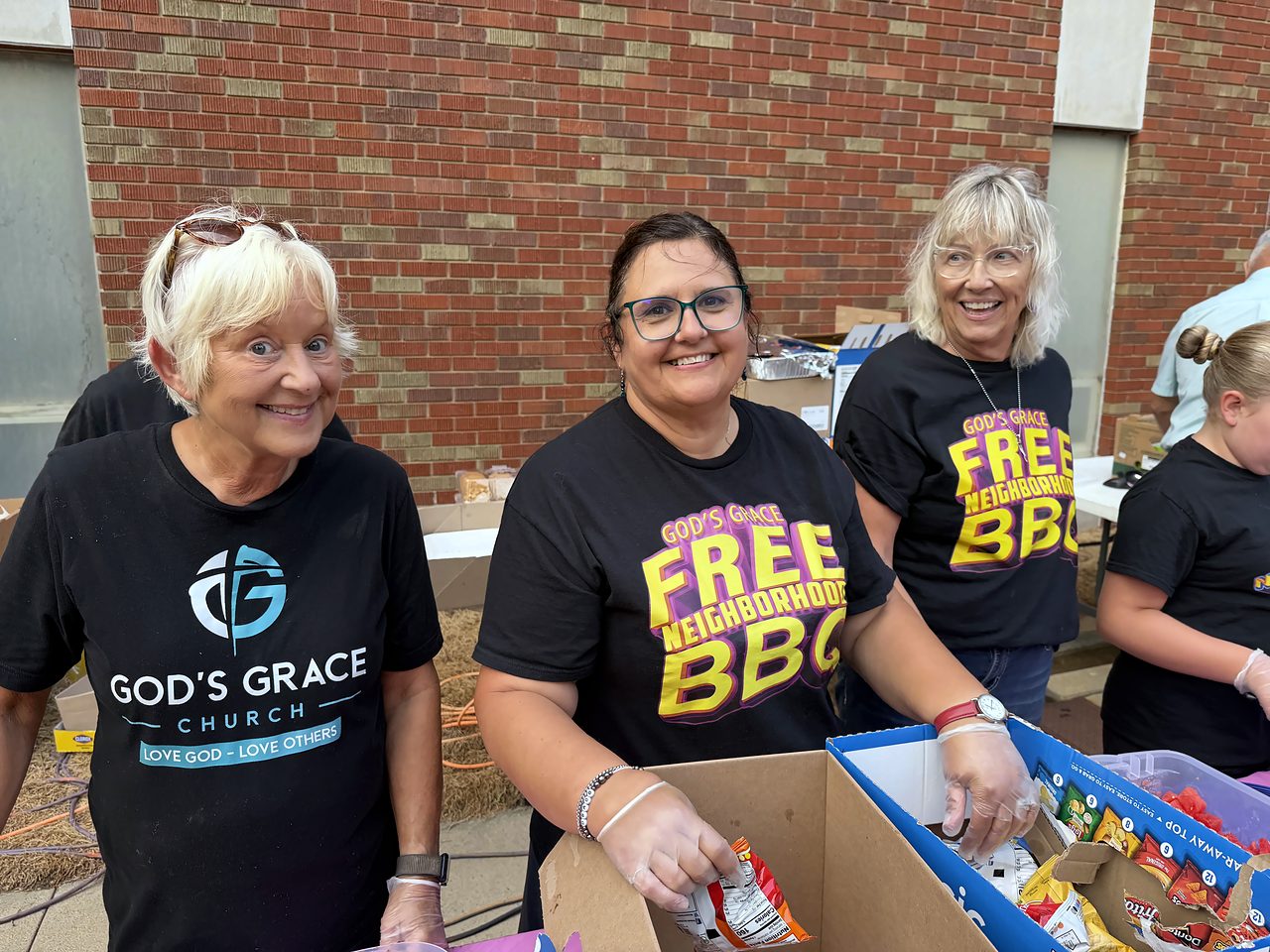 Smiling volunteers in God's Grace and Free Neighborhood BBQ shirts packing boxes